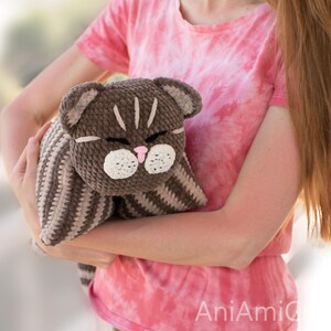May include: A brown and beige striped crochet cat plush toy with a dark brown head, white muzzle, pink nose, and closed eyes. The cat features stitched facial details and a soft, textured yarn construction, presented as a stuffed animal.