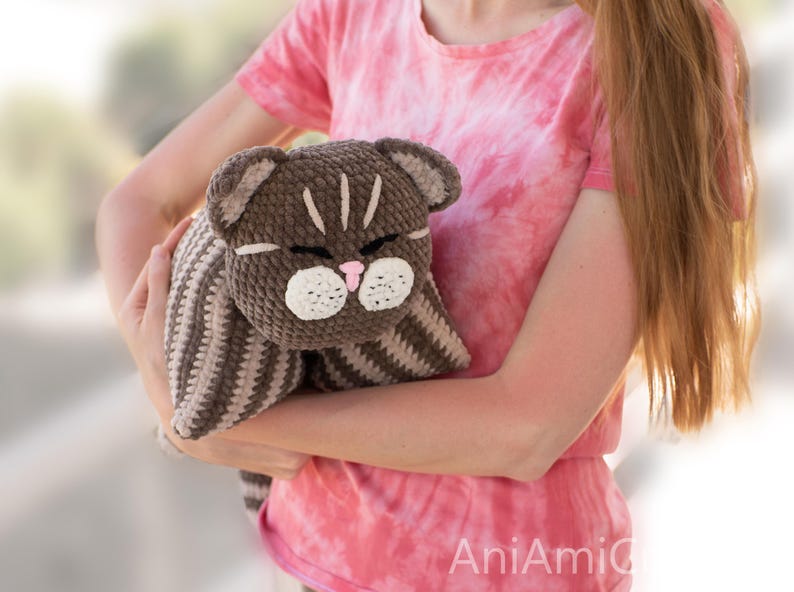 May include: A brown and beige striped crochet cat plush toy with a dark brown head, white muzzle, pink nose, and closed eyes. The cat features stitched facial details and a soft, textured yarn construction, presented as a stuffed animal.