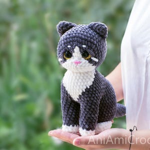 May include: A handmade crochet cat plush toy featuring dark gray and white yarn. The cat has large, glossy dark eyes with gold flecks, a small pink nose, and white markings on its chest and paws. The texture is soft and plush throughout.