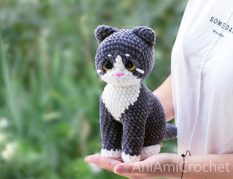 May include: A handmade crochet cat plush toy featuring dark gray and white yarn. The cat has large, glossy dark eyes with gold flecks, a small pink nose, and white markings on its chest and paws. The texture is soft and plush throughout.