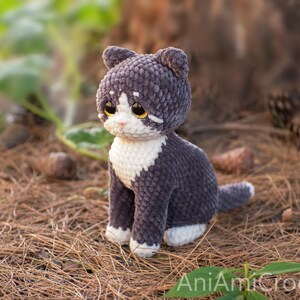 May include: A handmade crochet cat plush toy featuring dark gray and white yarn. The cat has large amber-colored eyes, a white muzzle, a white chest, and white paws. The text AniAmiCrochet is visible in the bottom right corner of the image.