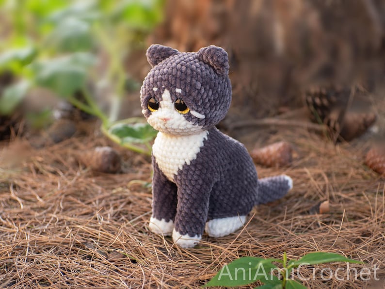 May include: A handmade crochet cat plush toy featuring dark gray and white yarn. The cat has large amber-colored eyes, a white muzzle, a white chest, and white paws. The text AniAmiCrochet is visible in the bottom right corner of the image.