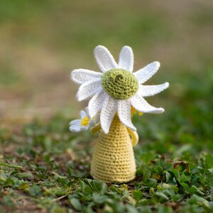 May include: A crocheted daisy doll with a white flower and yellow dress. The doll is sitting on a bed of green grass.