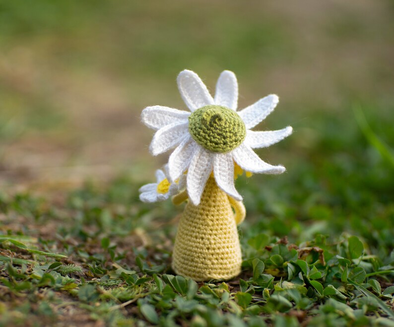 May include: A crocheted daisy doll with a white flower and yellow dress. The doll is sitting on a bed of green grass.