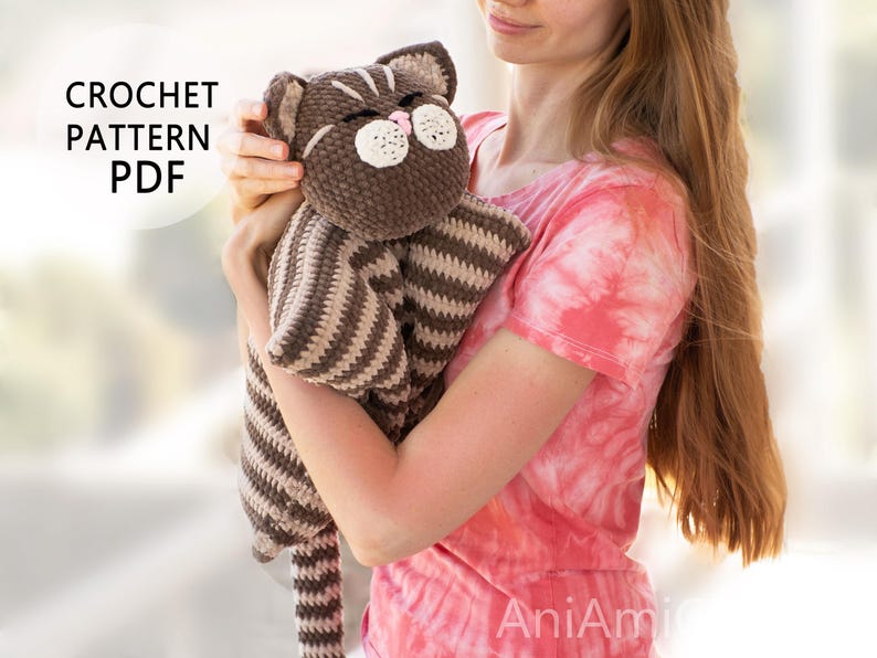 May include: A brown and beige striped crochet cat lovey with a plush head, embroidered eyes, and a pink nose. The text CROCHET PATTERN PDF is visible in the upper left corner. The cat has a soft, textured yarn finish and a long, striped tail.