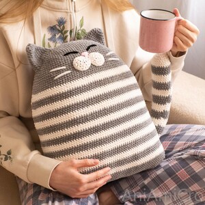 May include: A gray and white striped crocheted cat pillow with a pink nose and whiskers. The pillow is being held by a person sitting on a couch.