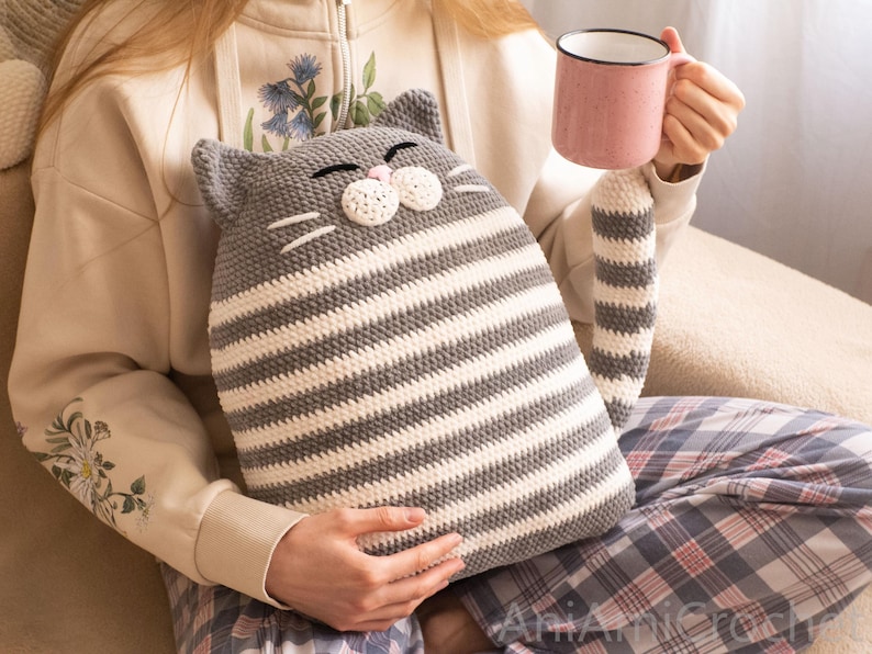 May include: A gray and white striped crocheted cat pillow with a pink nose and whiskers. The pillow is being held by a person sitting on a couch.