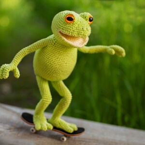 May include: A green crocheted frog toy riding a miniature skateboard. The frog has orange eyes and a toothy smile.