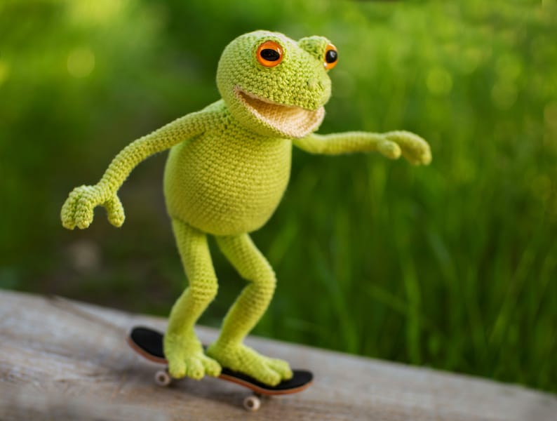 May include: A green crocheted frog toy riding a miniature skateboard. The frog has orange eyes and a toothy smile.