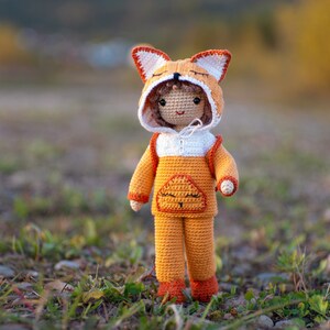 May include: A crocheted doll wearing an orange and white fox costume. The doll has a smiling face and is standing on a bed of green grass and brown rocks.