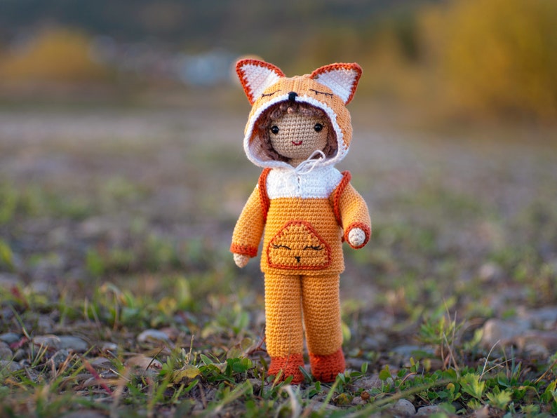 May include: A crocheted doll wearing an orange and white fox costume. The doll has a smiling face and is standing on a bed of green grass and brown rocks.