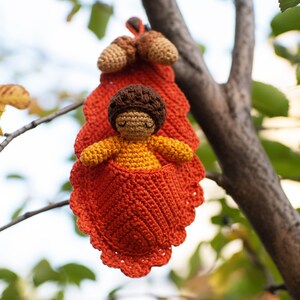 May include: A crocheted doll with brown hair and a yellow dress is sitting inside a crocheted orange leaf. The leaf is hanging from a tree branch.