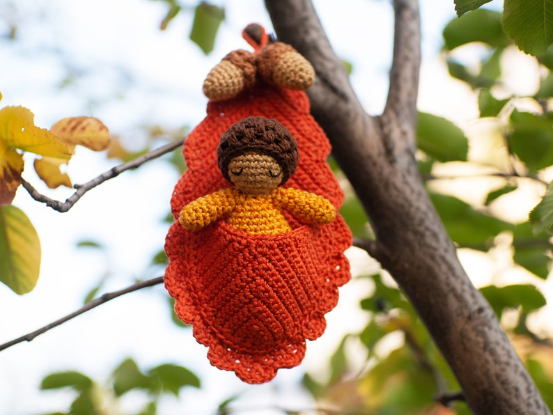 May include: A crocheted doll with brown hair and a yellow dress is sitting inside a crocheted orange leaf. The leaf is hanging from a tree branch.
