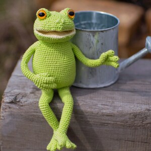 May include: A green crocheted frog toy sitting on a wooden surface next to a silver watering can.