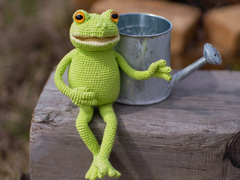 May include: A green crocheted frog toy sitting on a wooden surface next to a silver watering can.