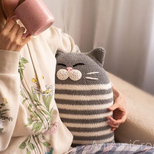 May include: A gray and white striped crocheted cat pillow with a pink nose and whiskers. The pillow is being held by a person wearing a floral patterned cream hoodie.