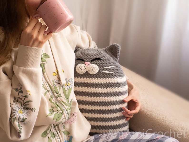 May include: A gray and white striped crocheted cat pillow with a pink nose and whiskers. The pillow is being held by a person wearing a floral patterned cream hoodie.