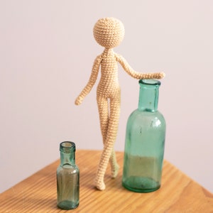 K&ouml;nnte beinhalten: Eine geh&auml;kelte Puppe mit einem beigefarbenen K&ouml;rper und einer gr&uuml;nen Glasflasche. Die Puppe steht auf einer Holzoberfl&auml;che.