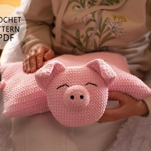 Op de afbeelding: Een lichtroze gehaakt varkensvormig kussen met zwarte ogen en een lachende mond. Het kussen heeft een getextureerd oppervlak en wordt door een persoon vastgehouden. De tekst "CROCHET PATTERN PDF" staat in een witte cirkel linksboven.