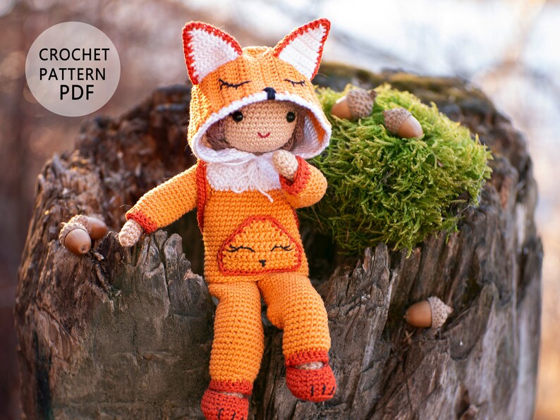 May include: A crocheted fox doll wearing a hooded fox costume, sitting on a tree stump with acorns. The doll has a white face with black eyes and a pink nose. The doll is wearing a crocheted orange and white outfit with a white fur trim. The doll is sitting on a tree stump with green moss and acorns.