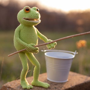 May include: A green crocheted frog toy holding a wooden stick, with a white bucket in the foreground. The frog has large, round, orange eyes and a wide smile.