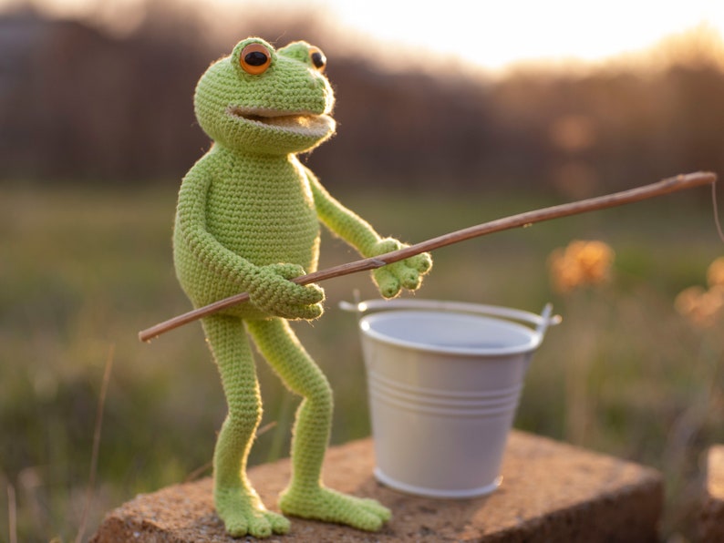May include: A green crocheted frog toy holding a wooden stick, with a white bucket in the foreground. The frog has large, round, orange eyes and a wide smile.