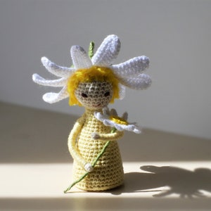 May include: A crocheted doll with yellow hair wearing a white daisy flower on its head and holding a yellow daisy flower in its hand.