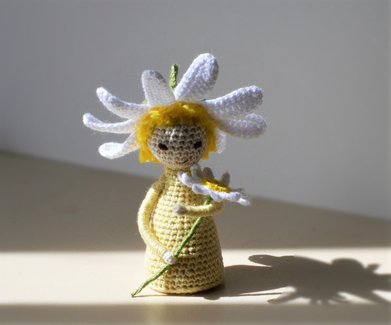 May include: A crocheted doll with yellow hair wearing a white daisy flower on its head and holding a yellow daisy flower in its hand.