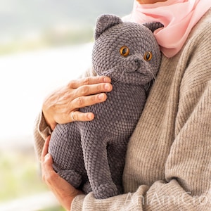 May include: A handmade gray crochet cat doll with orange eyes and whiskers. The plush cat is held by a person wearing a beige sweater and a pink scarf. The cat is a soft, cuddly toy.