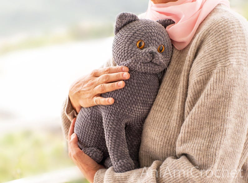 May include: A handmade gray crochet cat doll with orange eyes and whiskers. The plush cat is held by a person wearing a beige sweater and a pink scarf. The cat is a soft, cuddly toy.