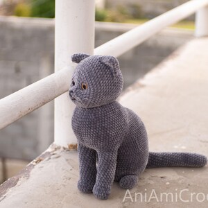 May include: A handmade, gray crocheted cat doll with yellow eyes. The plush cat is sitting upright with its tail extended. The doll is made of soft yarn and has a detailed face. The text "AniAmiCrochet" is visible in the bottom right corner.