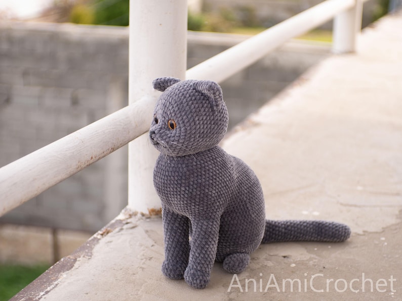 May include: A handmade, gray crocheted cat doll with yellow eyes. The plush cat is sitting upright with its tail extended. The doll is made of soft yarn and has a detailed face. The text "AniAmiCrochet" is visible in the bottom right corner.