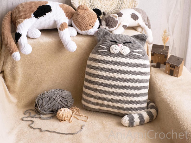 May include: A gray and white striped crocheted cat pillow with a white face and black eyes. The pillow is lying on a beige surface with two other crocheted cat toys.