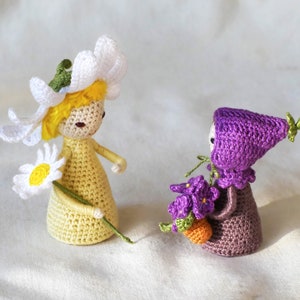 May include: Two crocheted dolls, one with a white daisy flower crown and a yellow dress holding a daisy, the other with a purple hat and dress holding a bouquet of purple flowers.