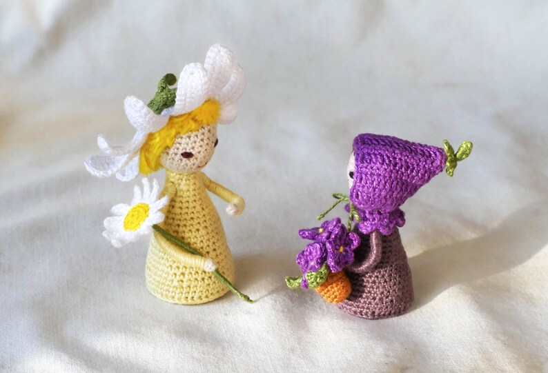 May include: Two crocheted dolls, one with a white daisy flower crown and a yellow dress holding a daisy, the other with a purple hat and dress holding a bouquet of purple flowers.