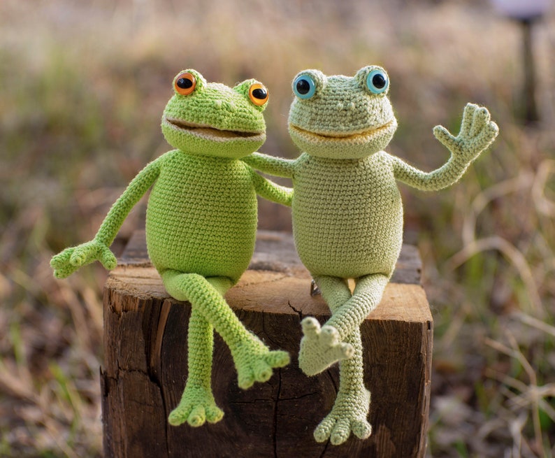 May include: Two crocheted green frog toys sitting on a tree stump. The frog on the left has orange eyes and the frog on the right has blue eyes.