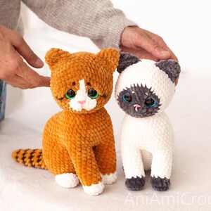 May include: Two plush cat toys made of soft yarn. One cat is orange with white paws and a white face patch, featuring large green eyes. The other cat is cream-coloured with dark grey ears, face, and paws, featuring large blue eyes. Text reads AniAmiCrochet.
