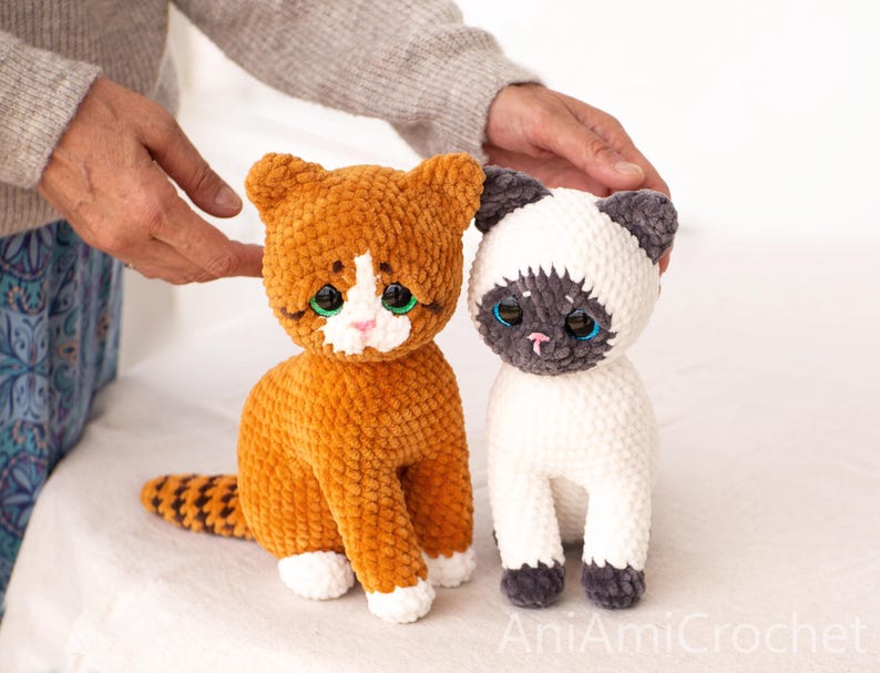 May include: Two plush cat toys made of soft yarn. One cat is orange with white paws and a white face patch, featuring large green eyes. The other cat is cream-coloured with dark grey ears, face, and paws, featuring large blue eyes. Text reads AniAmiCrochet.