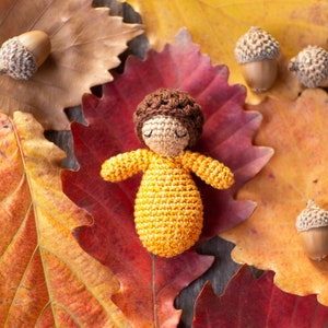 May include: A crocheted doll with brown hair and a yellow dress is lying on a bed of autumn leaves. The leaves are red, orange, and yellow.