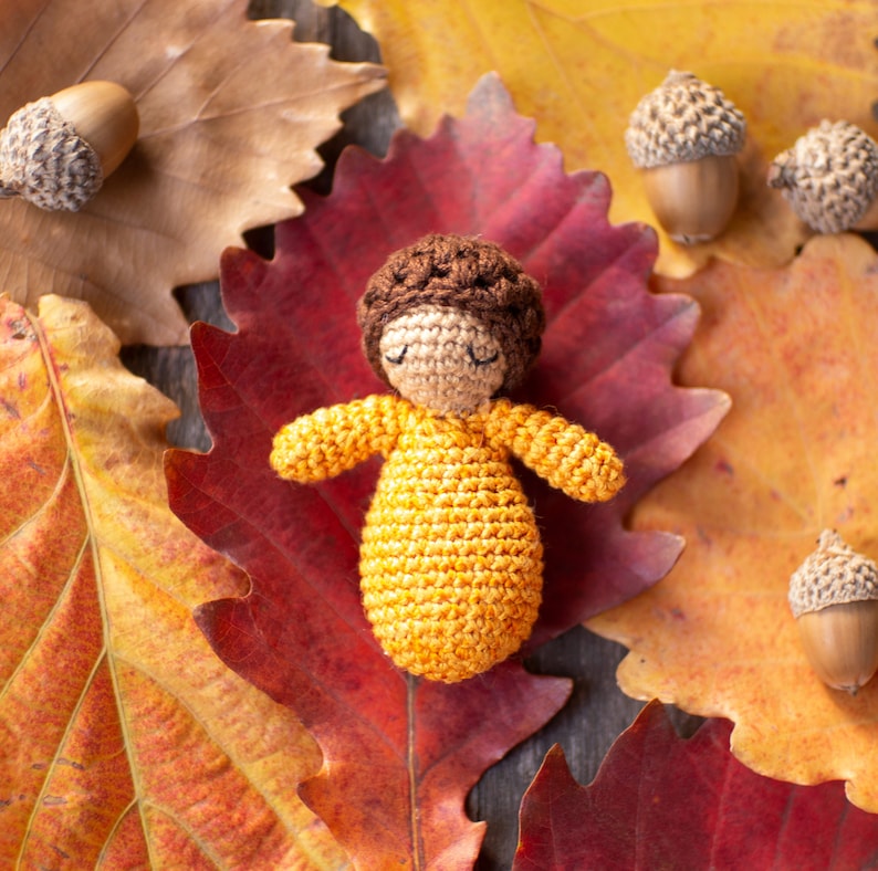 May include: A crocheted doll with brown hair and a yellow dress is lying on a bed of autumn leaves. The leaves are red, orange, and yellow.