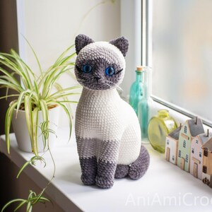 May include: A crocheted Siamese cat plush toy with a cream-colored body, dark gray ears, face, and paws, and bright blue plastic eyes. The cat is sitting upright and features the text AniAmiCrochet in the bottom right corner of the image.