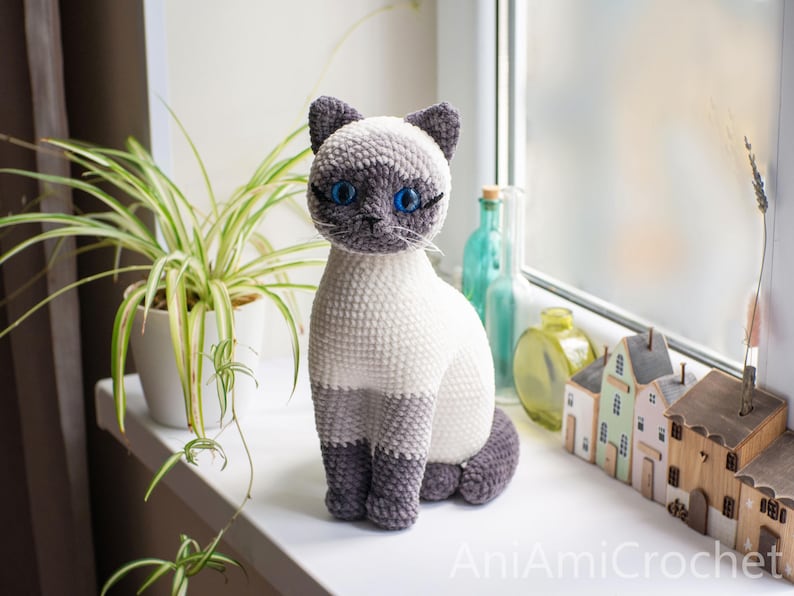 May include: A crocheted Siamese cat plush toy with a cream-colored body, dark gray ears, face, and paws, and bright blue plastic eyes. The cat is sitting upright and features the text AniAmiCrochet in the bottom right corner of the image.