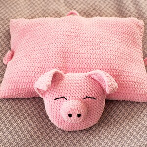 K&ouml;nnte beinhalten: Ein rechteckiges, geh&auml;keltes Kissen in Rosa mit einem angebrachten Pl&uuml;sch-Schweinekopf auf der Vorderseite. Der Schweinekopf hat zwei Schlappohren, eine Schnauze mit zwei schwarzen Nasenl&ouml;chern und mit schwarzem Garn gestickte, geschlossene Augen.