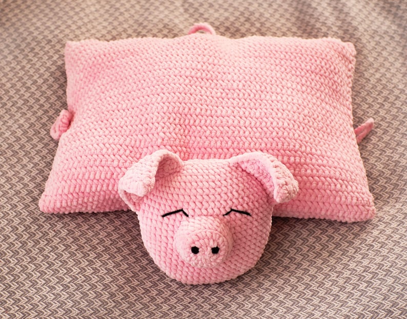 K&ouml;nnte beinhalten: Ein rechteckiges, geh&auml;keltes Kissen in Rosa mit einem angebrachten Pl&uuml;sch-Schweinekopf auf der Vorderseite. Der Schweinekopf hat zwei Schlappohren, eine Schnauze mit zwei schwarzen Nasenl&ouml;chern und mit schwarzem Garn gestickte, geschlossene Augen.