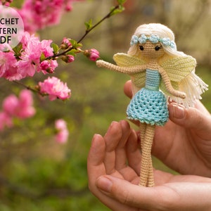 May include: Crochet pattern PDF for a small, light blue and yellow fairy doll with long blonde hair and a flower crown. The doll is holding a pink flower in its hand.