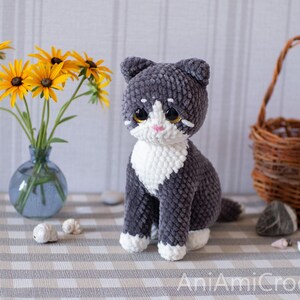 May include: A handmade crochet cat toy featuring dark gray and white yarn. The cat has large, dark, glossy eyes, a small pink nose, and white markings on its face, chest, and paws. The text AniAmiCrochet is visible in the bottom right corner.