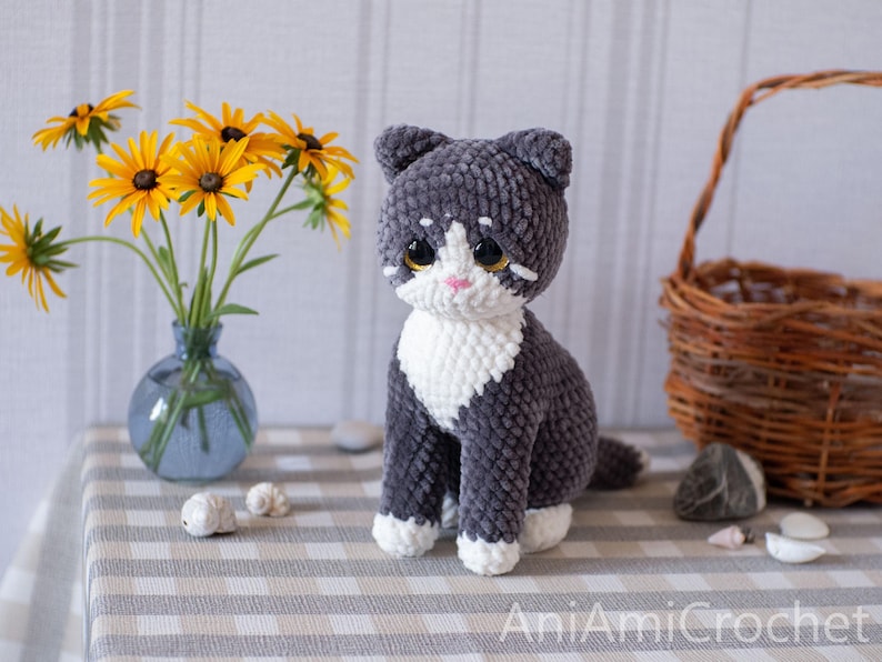 May include: A handmade crochet cat toy featuring dark gray and white yarn. The cat has large, dark, glossy eyes, a small pink nose, and white markings on its face, chest, and paws. The text AniAmiCrochet is visible in the bottom right corner.