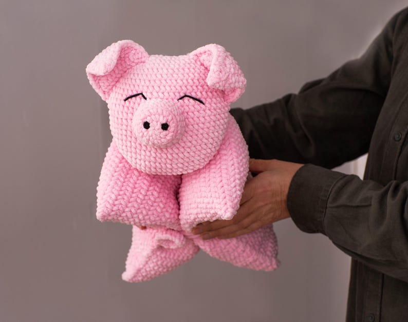 K&ouml;nnte beinhalten: Ein handgeh&auml;keltes Schweinchen-Stofftier aus weichem, hellrosa Garn. Das Kuscheltier hat einen runden Kopf mit gro&szlig;en, h&auml;ngenden Ohren, eine ausgepr&auml;gte Schnauze mit zwei schwarzen Nasenl&ouml;chern und mit schwarzem Faden gestickte, geschlossene Augen.