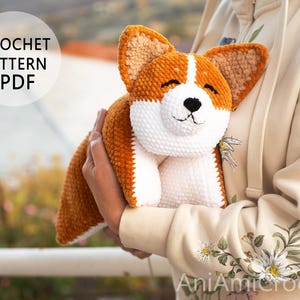 May include: A crocheted corgi dog plushie in shades of orange, white, and brown. The dog has a smiling face with black eyes and nose. The image includes the text "CROCHET PATTERN PDF".