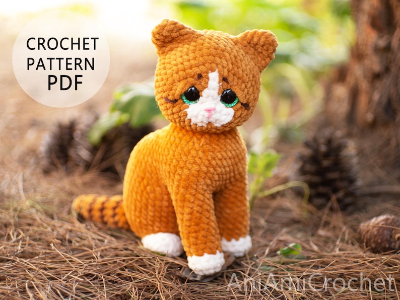 May include: A crocheted ginger cat toy with white paws, a white chest, and a white muzzle. The cat has large, dark, glossy eyes with green accents and a small pink nose. Text in the upper left corner reads CROCHET PATTERN PDF.